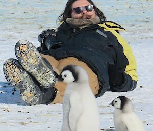 A man lies on ice near some back Emperor chicks.