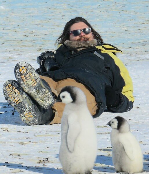 A man lies on ice near some back Emperor chicks.