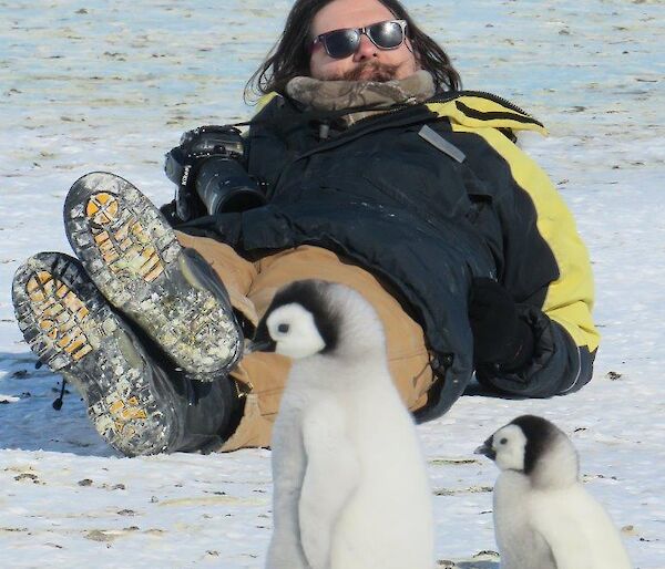 A man lies on ice near some back Emperor chicks.