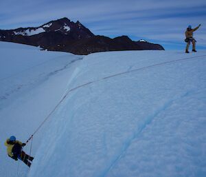 One man over a cliff on a rope while another watches