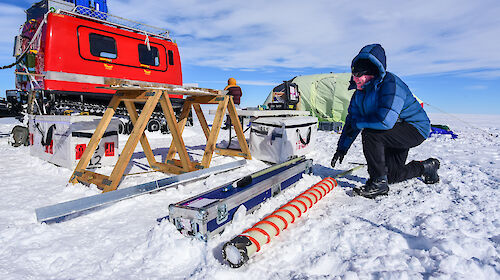 A scientist extracts ice from the ice core drill, with the campsite and Hägglunds in the background.