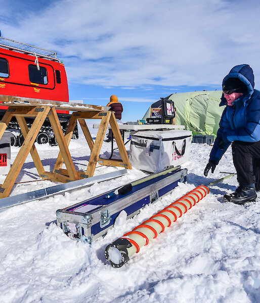 A scientist extracts ice from the ice core drill, with the campsite and Hägglunds in the background.