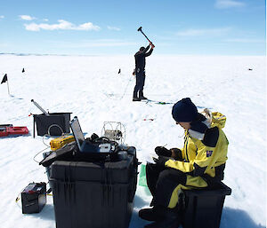 One expeditioner brings a hammer down onto a metal plate on the glacier while another expeditioner collects data from the hammer strike, relayed to a laptop computer.