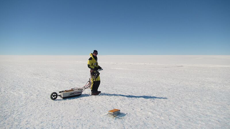 Tom hauls a ground penetrating radar antenna in a box with a small wheel across the ice. Tom is roped into a harness to allow him to haul the box behind him.