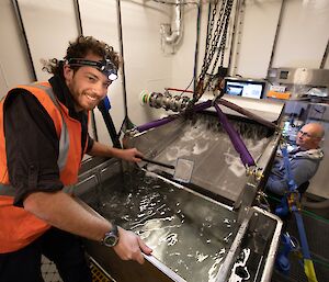 Two scientists inside a watertight room on Nuyina, with one standing at a steel filter table monitoring a flow of water across it.