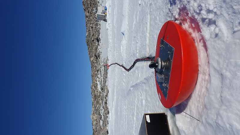 A modified sea ice buoy painted red, sits on top of the sea ice and carries electronic cables and water pipes below the ice