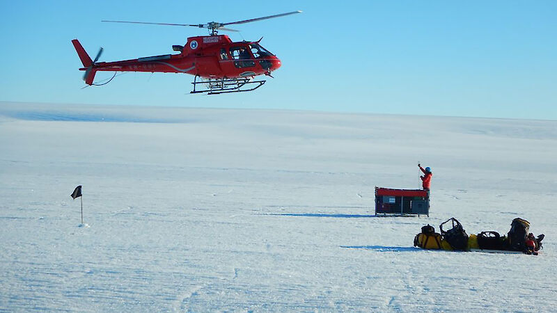 Red helicopter hovering over glacier surface as it maneuvers to pick up sling load