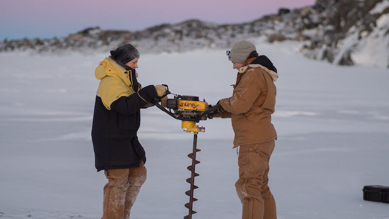 2 expeditioners hold a large drill on the ice