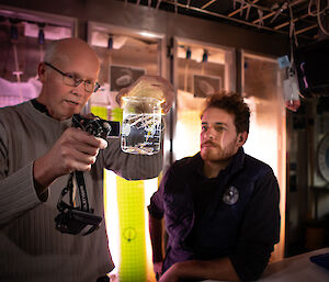 Two scientists in a darkened aquarium lab look at a beaker full of water and krill specimens, lit by a torch.
