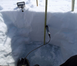 In this 49cm-deep snow pit the sea ice surface is visible at the bottom. The photo shows a probe measuring snow temperature, and a metal cutter of known volume used to acquire snow density samples up the snow column.