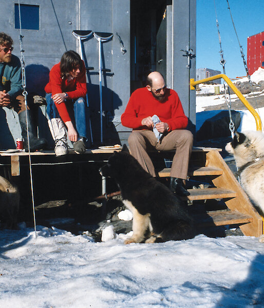 Tom sitting with other expeditioners on the steps outside a building at Mawson with huskies.