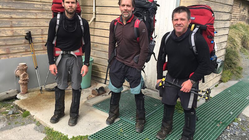 The Macquarie Island field hut assessment team of Paul Farrow (left), Dr Aleks Terauds and Ben Wood from VEC Civil Engineering (right), dressed in hiking gear and packs, outside a hut.