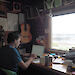 Aleks Terauds sitting at a laptop on a desk inside a field hut with a view to outside and various books, a guitar and tea pot surrounding him.