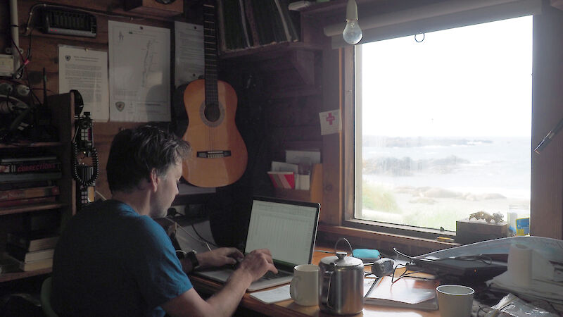 Aleks Terauds sitting at a laptop on a desk inside a field hut with a view to outside and various books, a guitar and tea pot surrounding him.