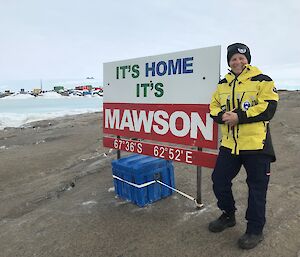 An expeditioner stands next to a sign that says "It's home, it's Mawson".