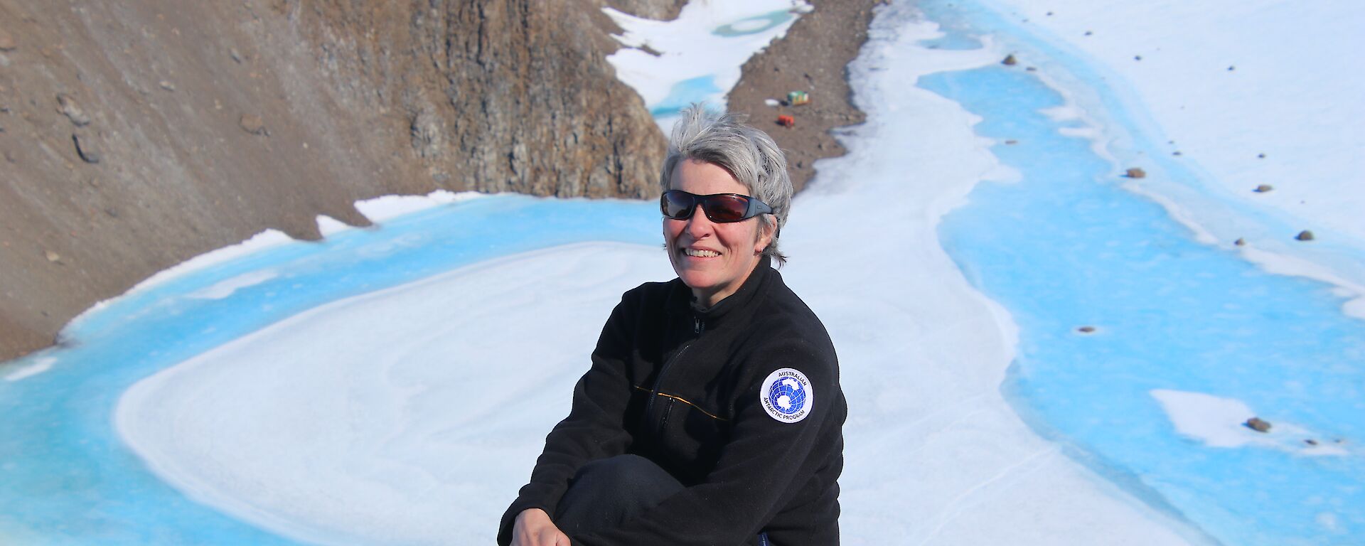 An expeditioner sits on a high, rocky outcrop. Below, white ice and blue melt water meed rocky ground. There are a couple of colourful buildings in the distance.