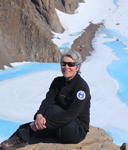 An expeditioner sits on a high, rocky outcrop. Below, white ice and blue melt water meed rocky ground. There are a couple of colourful buildings in the distance.