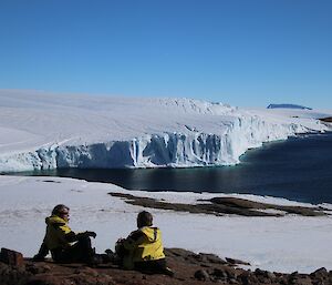 2 expeditioners sit on rocky ground overlooking a bay with ice cliffs.