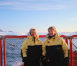 2 expeditioners stand on the deck of a ship with an icy landscape in the background.