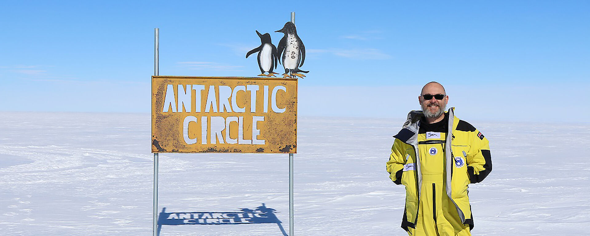 An expeditioner stands next to a sign that says "Antarctic Circle".