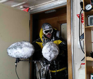 A snow-covered expeditioner holding 2 fluffy microphones enters through a door.