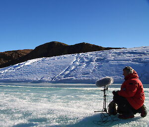 An expeditioner in red kneels on a frozen body of water with a fluffy microphone.