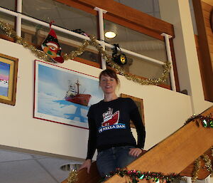 An artist stands on a staircase, next to painting of a ship - the Nella Dan. She is wearing a t-shirt with a picture of the same ship on it.