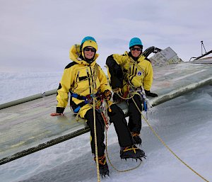 Two expeditioners sitting on wing of wrecked plane in snow.