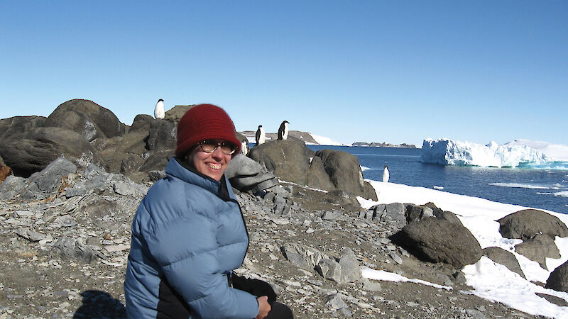 Researcher kneeling on the rocky shoreline with Adelie penguins in background