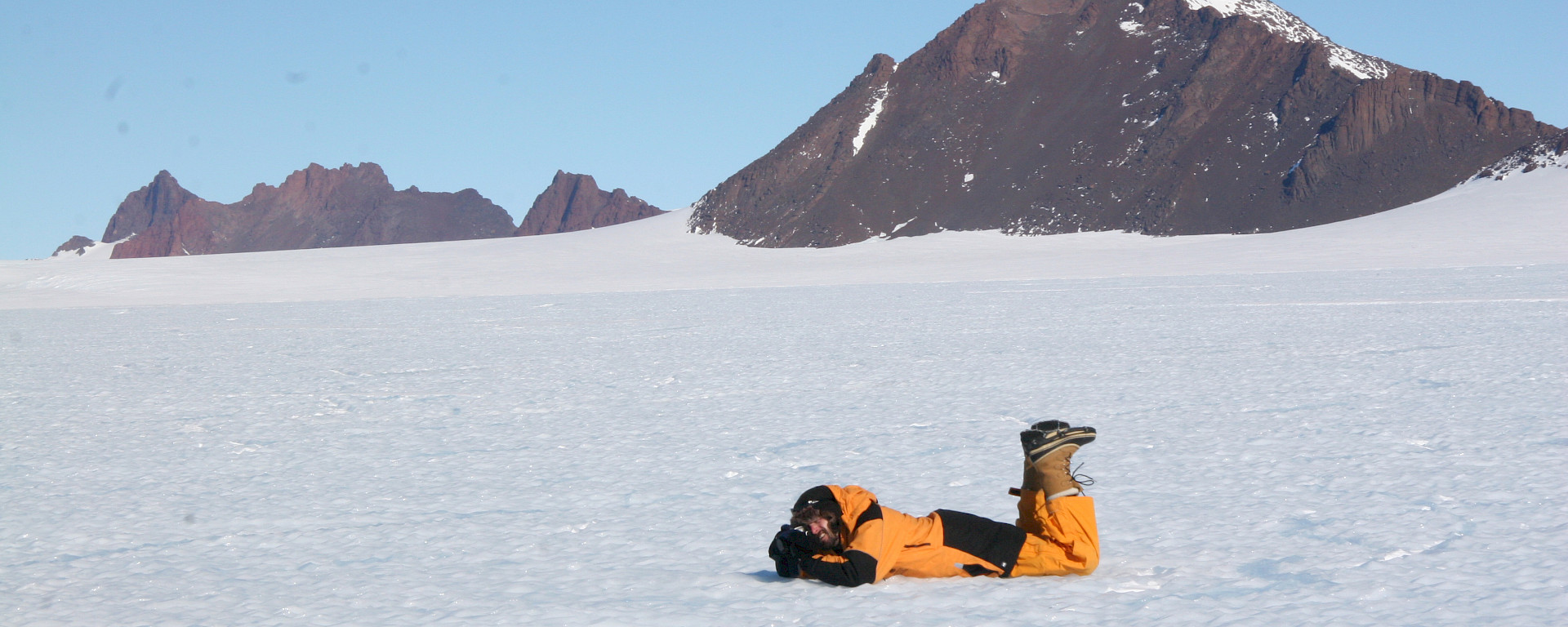 Nick Hutcheson lies on his stomach in the snow in Antarctica, camera in hand, aiming at an unseen subject