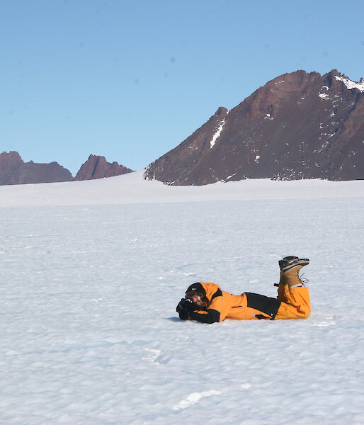 Nick Hutcheson lies on his stomach in the snow in Antarctica, camera in hand, aiming at an unseen subject