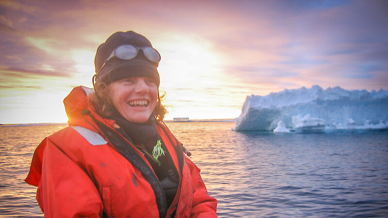 An expeditioner dressed in red sits in a small boat in front of an iceberg.