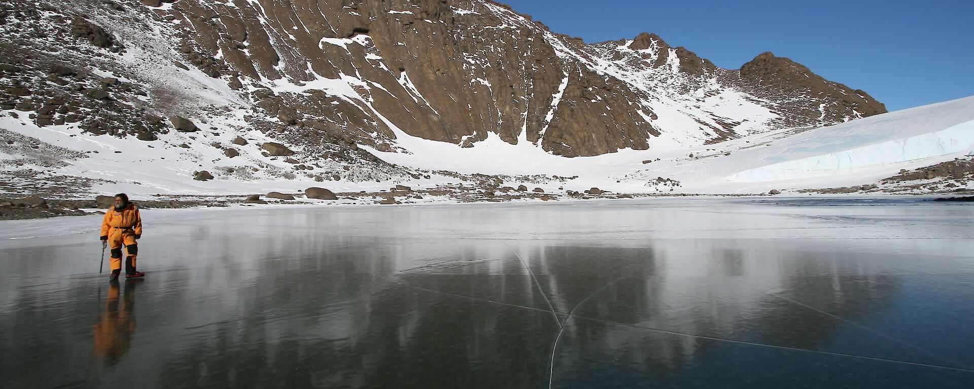 An expeditioner stands on a frozen lake with a rocky hill behind.