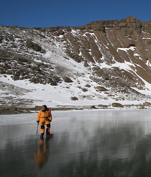 An expeditioner stands on a frozen lake with a rocky hill behind.