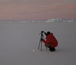 An expeditioner kneels on the ice with a camera on a tripod.