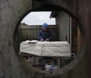 Looking through oval cut out in fence to meteorologist using large bench mounted equipment outdoors