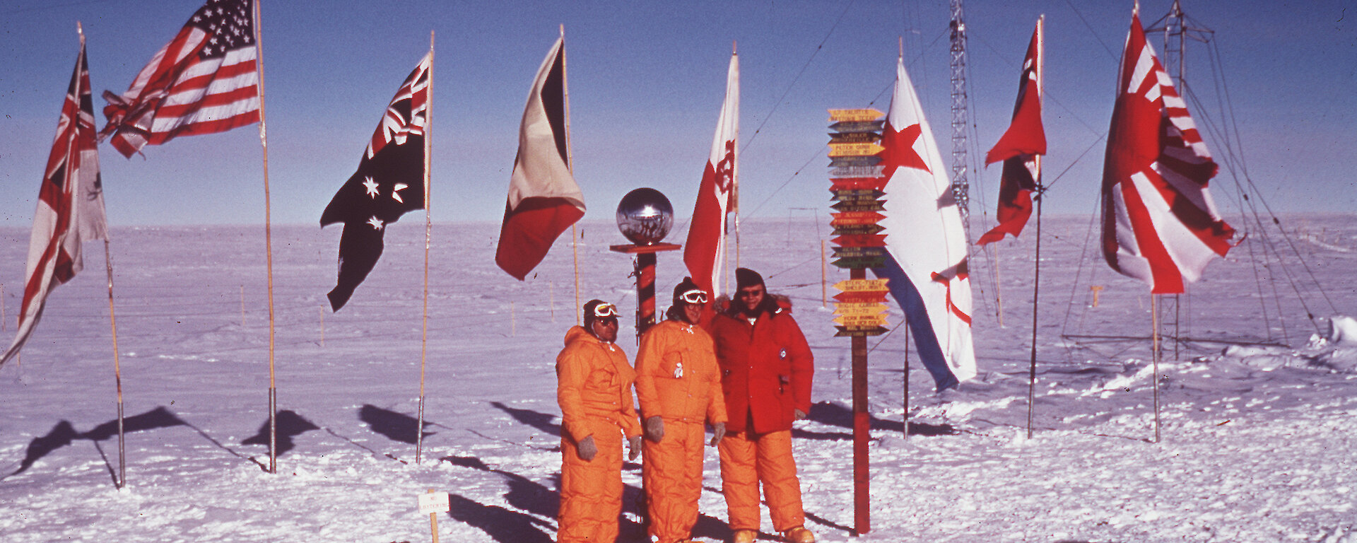 Three men standing at South Pole marker with row of international flags behind