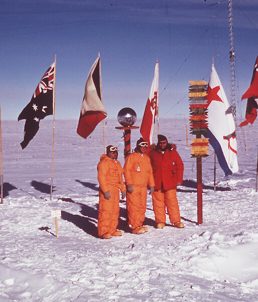 Three men standing at South Pole marker with row of international flags behind