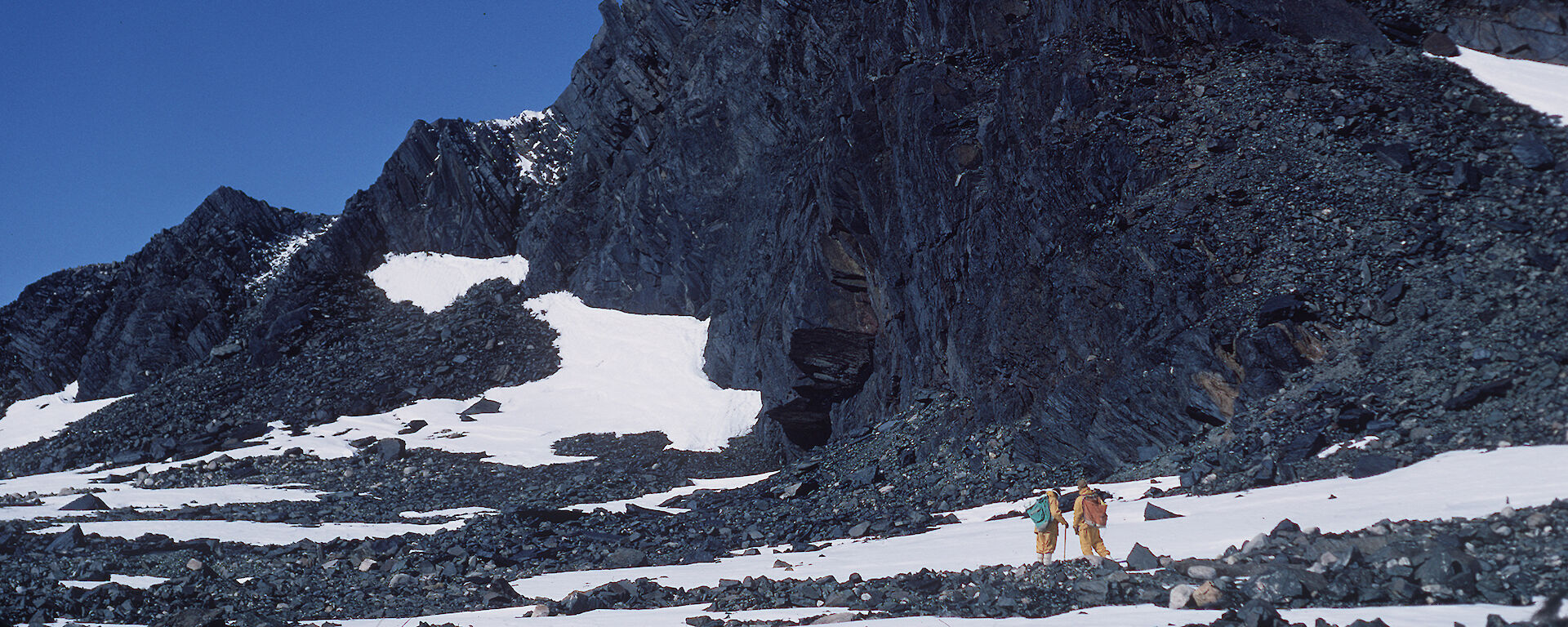 Expeditioners walk beneath a dark rocky outcrop.