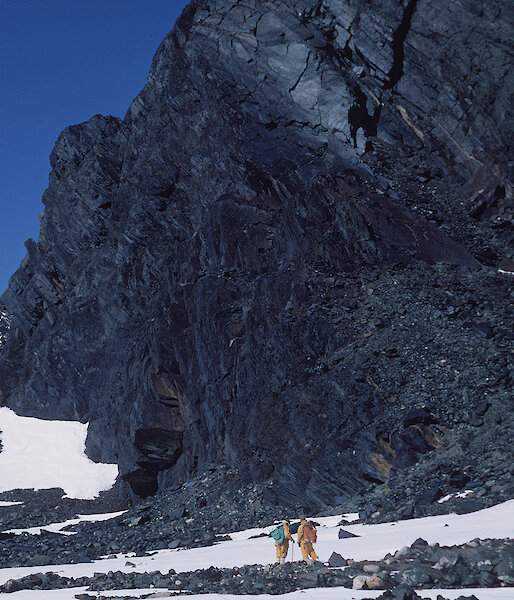 Expeditioners walk beneath a dark rocky outcrop.