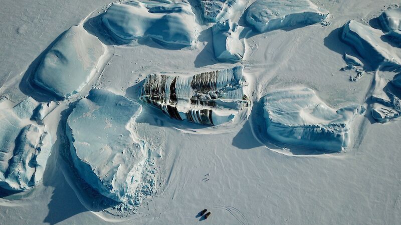 Several icebergs including a striped iceberg on the sea ice with a small group of expeditioners just visible and some Hägglunds tracks