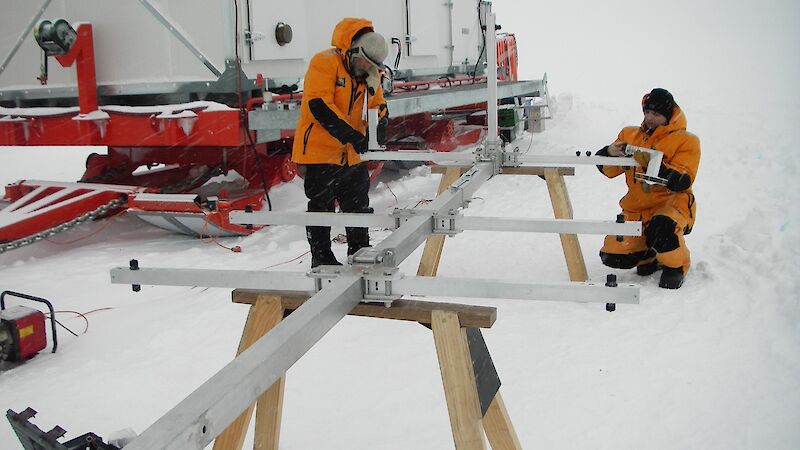 Long metal mast with metals stays in laid out across saw horses in the snow during assembly.