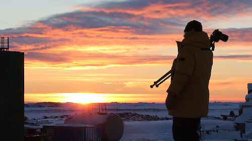 Expeditioner with camera tripod over shoulder walks in front of rising sun