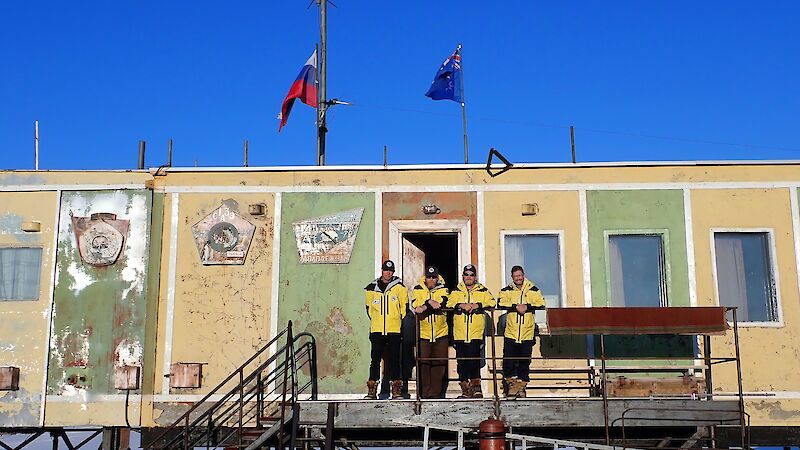 Four men wearing cold weather clothing standing on the balcony of a square building.