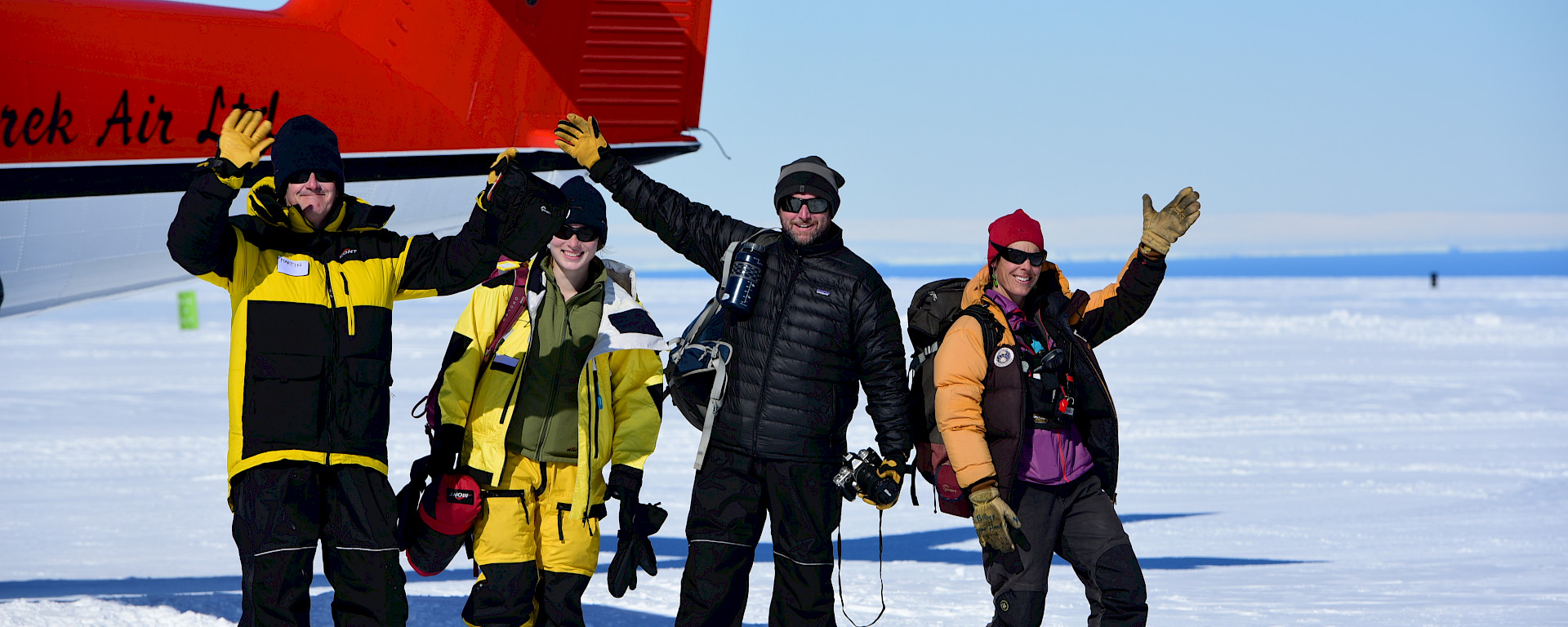 Four happily waving expeditioners, near the tail of the aircraft, on the white ski landing area