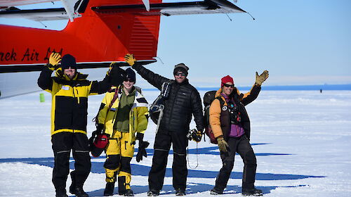 Four happily waving expeditioners, near the tail of the aircraft, on the white ski landing area