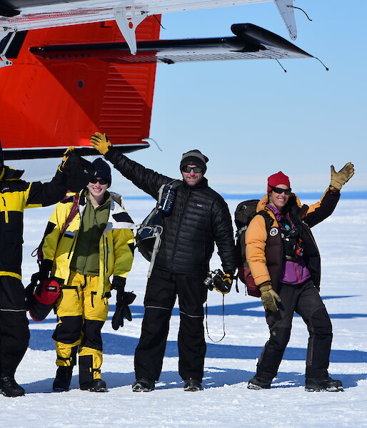 Four happily waving expeditioners, near the tail of the aircraft, on the white ski landing area