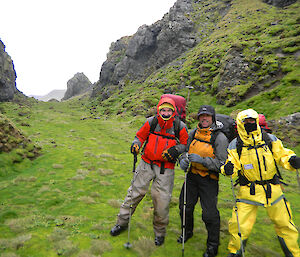 Three survival suit dressed men walking between high tall rock faces with lush greenery on ground
