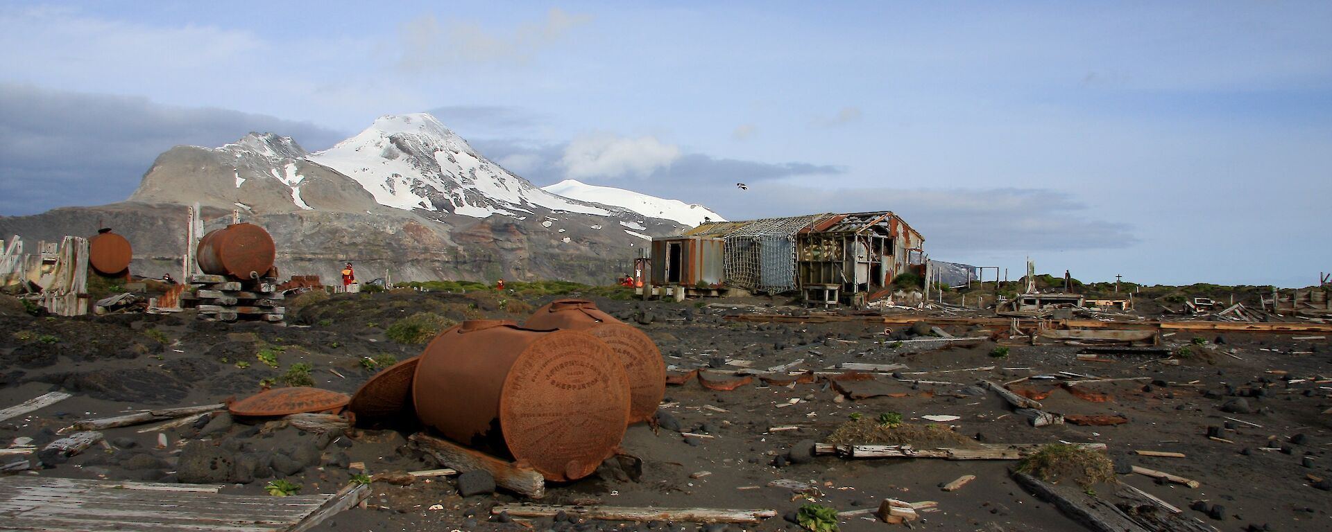 Abandoned huts and rusted tanks at former station site with snowcapped hill in background