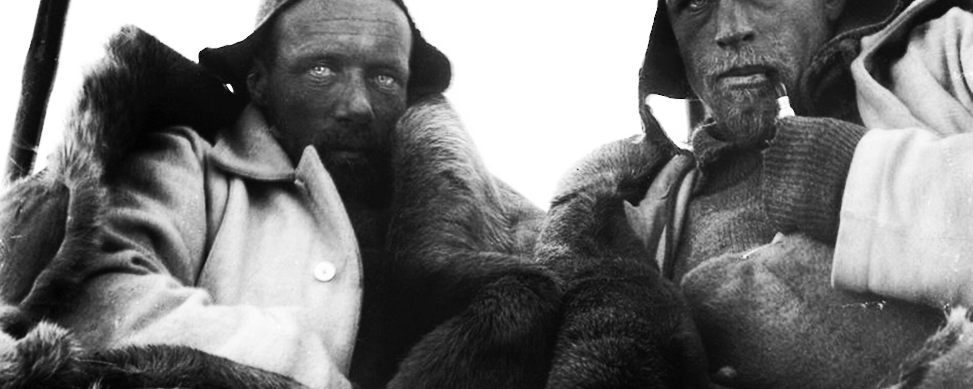 Two men wearing woollen beanies and hunching to keep warm stare at the camera with weathered faces.